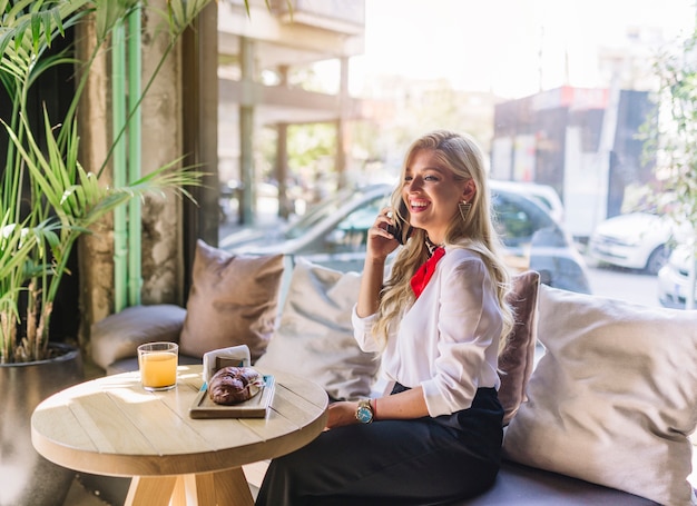 Happy young woman talking on mobile phone with baked bread and juice on table