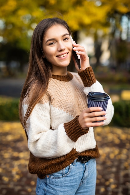 Felice giovane donna parla al telefono, tenendo una tazza di caffè da asporto e sorridente.