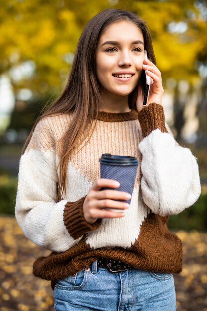 電話で話している、持ち帰り用のコーヒーカップを押しながら笑って幸せな若い女。