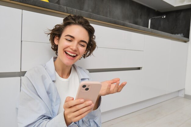 Happy young woman talks on mobile phone video chats on smartphone app sits on floor at home