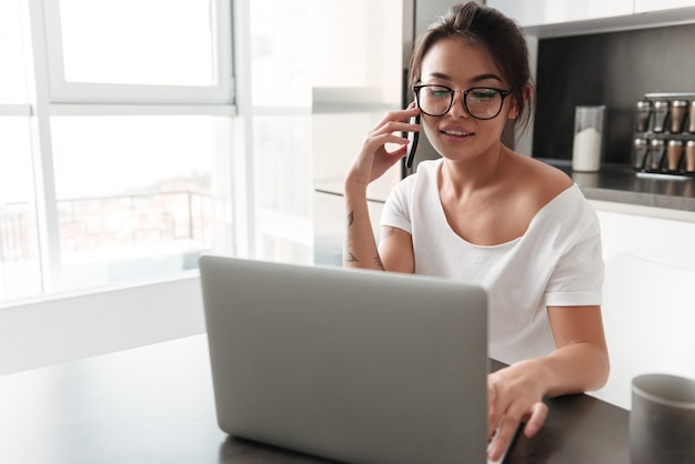 Happy young woman using laptop talking by mobile phone.