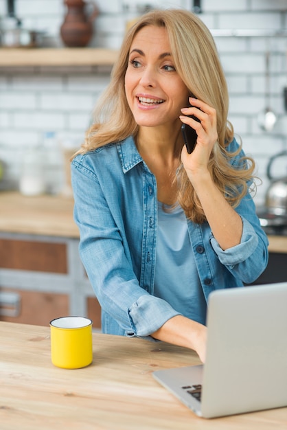 Happy young woman using laptop talking on smart phone in the kitchen