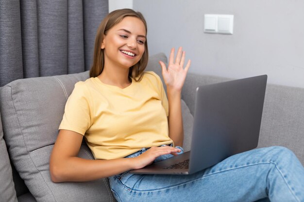 Happy young woman video calling sitting on sofa and using laptop at home