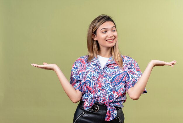 A happy young woman wearing paisley printed shirt smiling and raising hands on a green wall