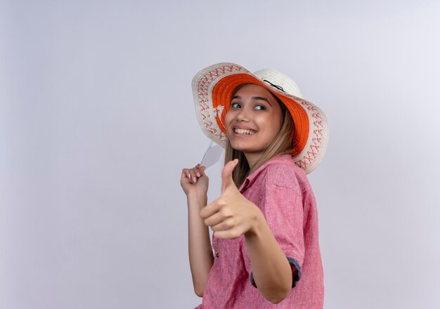 Счастливая молодая женщина в красной рубашке держит билеты на самолет с большими пальцами руки вверх, глядя в сторону на белой стене