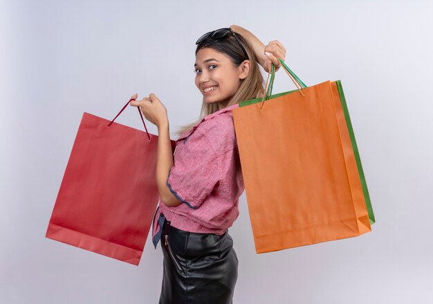 A happy young woman wearing red shirt showing colorful shopping bags while looking on a white wall