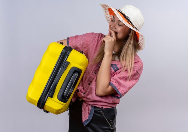 A happy young woman wearing red shirt and sun hat keeping hand on mouth while carrying a yellow suitcase on a white wall