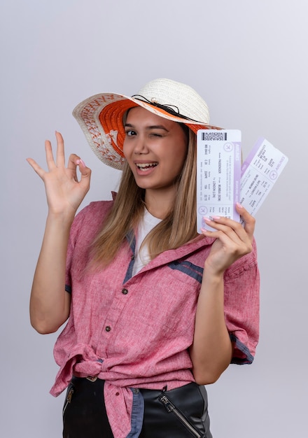 흰 벽에 확인 제스처와 함께 비행기 티켓을 보여주는 빨간 셔츠와 태양 모자를 쓰고 행복 한 젊은 여자