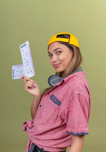A happy young woman wearing red shirt and yellow baseball hat showing plane tickets while looking on a green wall