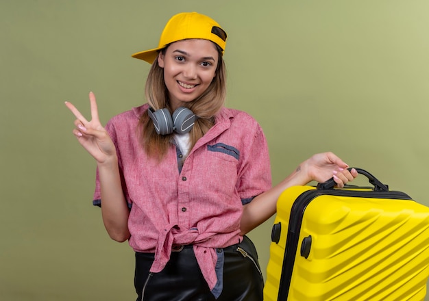 Счастливая молодая женщина в красной рубашке и желтой бейсболке показывает жест двумя пальцами, неся желтый чемодан на зеленой стене