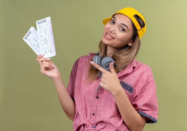 A happy young woman wearing red shirt and yellow baseball hat with headphones pointing at plane tickets with index finger on a green wall