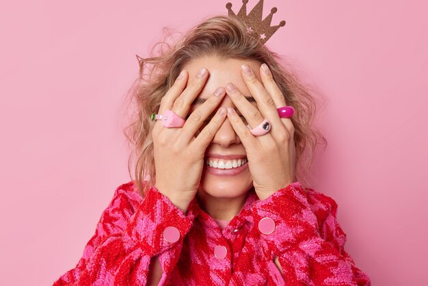 Happy young woman wears crown and jacket covers eyes with hands awaits for surprise smils broadly shows white teeth prepares for celebration isolated over pink background going to get present