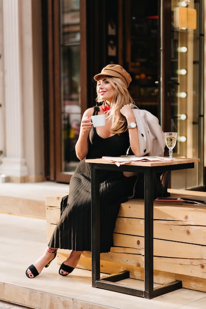 Happy young woman wears trendy black shoes and long dress relaxing after hard day and drinking coffee. Outdoor portrait of smiling girl in brown cap and coat waiting friend to celebrate something.