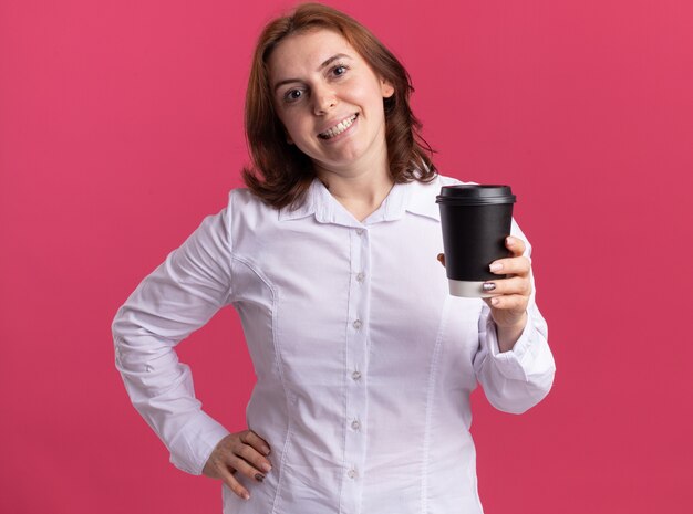 Счастливая молодая женщина в белой рубашке держит чашку кофе, глядя вперед, весело улыбаясь, стоя над розовой стеной