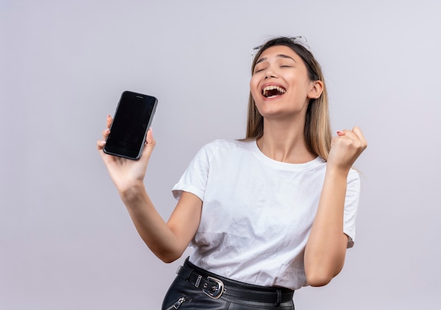 Счастливая молодая женщина в белой футболке в солнечных очках улыбается, показывая пустое место мобильного телефона со сжатым кулаком на белой стене