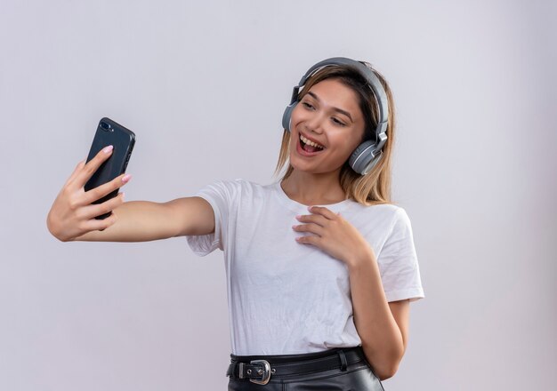 白い壁にスマートフォンでselfieを撮るヘッドフォンを身に着けている白いTシャツの幸せな若い女性