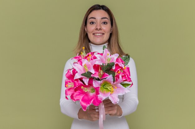 Счастливая молодая женщина в белой водолазке держит букет цветов, глядя вперед, весело улыбаясь, празднует международный женский день, стоя над зеленой стеной