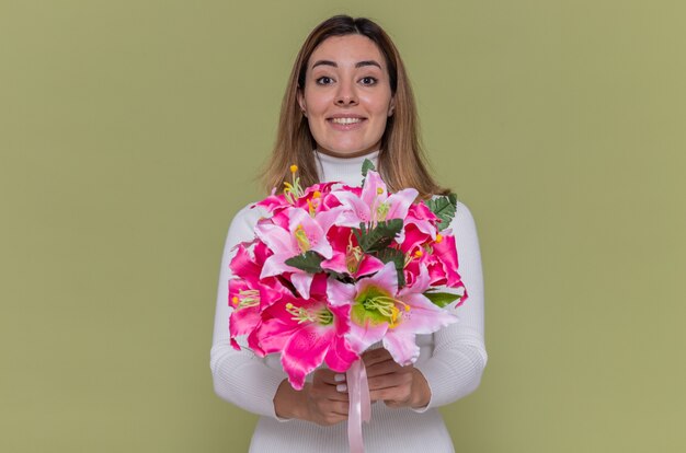 Happy young woman in white turtleneck holding bouquet of flowers looking at front smiling cheerfully celebrating international women's day standing over green wall