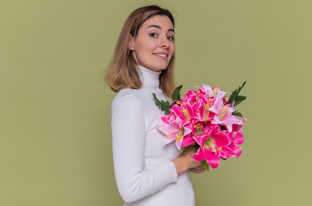 Happy young woman in white turtleneck holding bouquet of flowers looking at front smiling cheerfully celebrating international women's day standing over green wall