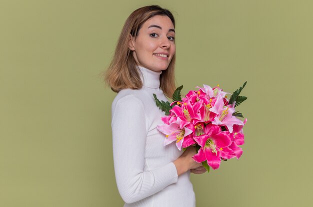 Happy young woman in white turtleneck holding bouquet of flowers looking at front smiling cheerfully celebrating international women's day standing over green wall