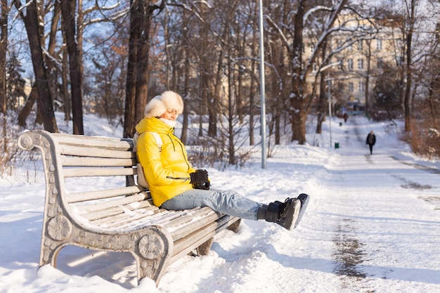 Счастливая молодая женщина зимой в теплой одежде в заснеженном парке в солнечный день сидит на скамейках и наслаждается свежим воздухом и кофе в одиночестве