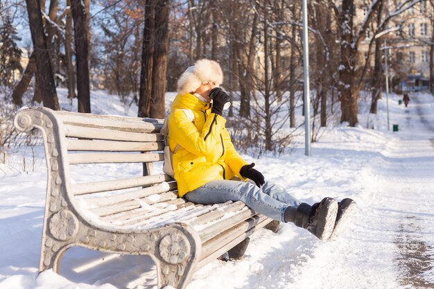 Happy young woman in winter in warm clothes in a snowy park on a sunny day sits on the benches and enjoys the fresh air and coffee alone