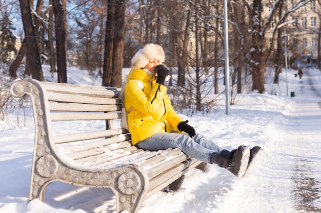 晴れた日に雪の公園で暖かい服を着て冬の幸せな若い女性はベンチに座って、新鮮な空気とコーヒーを一人で楽しんでいます