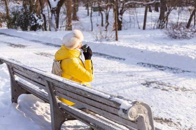 Happy young woman in winter in warm clothes in a snowy park on a sunny day sits on the benches and enjoys the fresh air and coffee alone