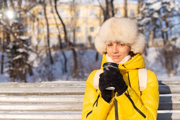 晴れた日に雪の公園で暖かい服を着て冬の幸せな若い女性はベンチに座って、新鮮な空気とコーヒーを一人で楽しんでいます