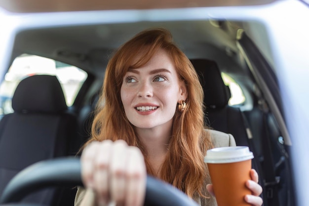 Счастливая молодая женщина с кофе за рулем своего автомобиля Женщина пьет кофе за рулем автомобиля Молодая женщина пьет кофе за рулем автомобиля Привлекательные рыжие волосы водят машину