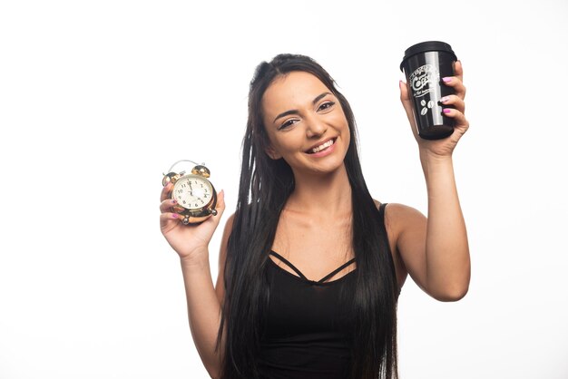 Happy young woman with cup holding an alarm clock on white wall