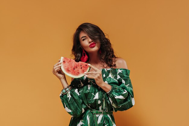 Happy young woman with curly dark hair and red lipstick in earrings and printed green dress posing with watermelon on orange wall