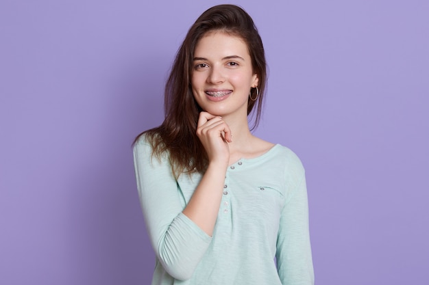 Happy young woman with dark hair keeping fist under chin, looks at camera, wears casual attire