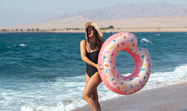 海のそばにドーナツ型の水泳サークルを持つ幸せな若い女性。休暇中のレジャーとエンターテイメントの概念。