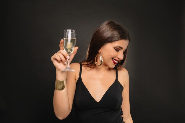 Happy young woman with golden jewerly in black dress drinking champagne