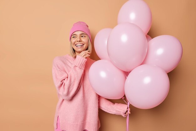 Happy young woman with joyful expression touches face gently wears hat and casual jumper poses with bunch of inflated balloons comes on party isolated over beige background. Holidays concept