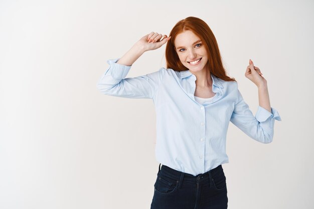 긴 빨간 머리를 가진 행복한 젊은 여성이 재미 있고, 춤을 추고, 일하는 하루를 즐기고, 흰 벽 위에 서 있습니다.