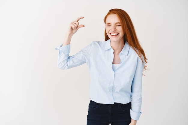 Happy young woman with long red hair showing something small, demonstrate tiny object and laughing, standing over white wall