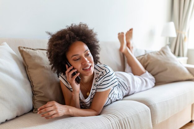 携帯電話がソファに横たわっている幸せな若い女性タブレットコンピューターを使用して美しい若い黒人女性