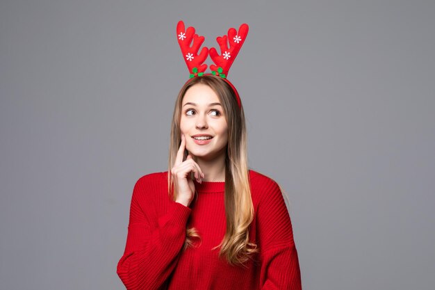 Happy young woman with reindeer horns on her head looks and pointing at camera on gray wall