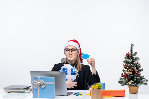 Free photo happy young woman with santa claus hat and wearing eyeglasses sitting at a table holding christmas gift and bank card on white background