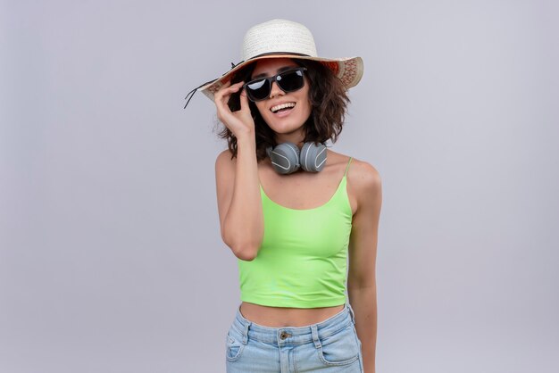 A happy young woman with short hair in green crop top in headphones wearing sunglasses and sun hat on a white background
