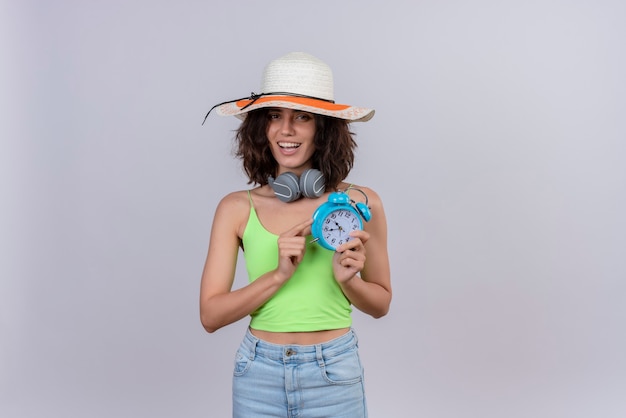 A happy young woman with short hair in green crop top wearing sun hat holding blue alarm clock on a white background