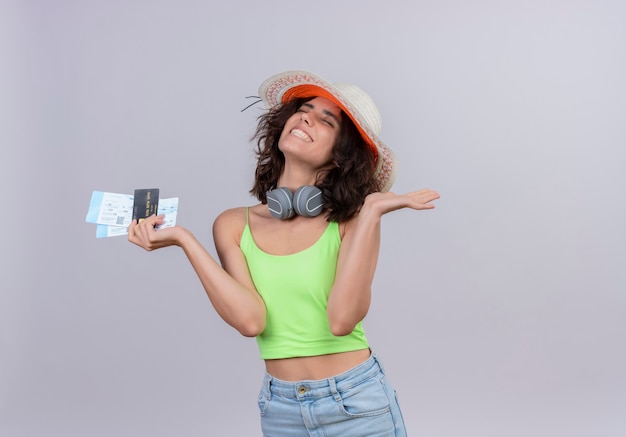 A happy young woman with short hair in green crop top wearing sun hat holding plane tickets and credit card on a white background
