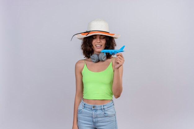 A happy young woman with short hair in green crop top wearing sun hat looking at a blue toy airplane on a white background