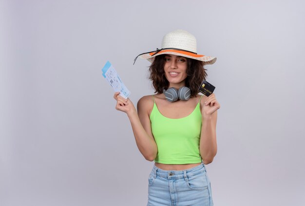 A happy young woman with short hair in green crop top wearing sun hat showing plane tickets and credit card on a white background