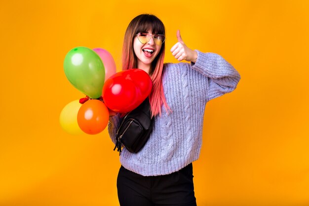 Happy young woman with unusual pink hairs having fun and posing at yellow wall, holding colorful birthday party balloons, casual trendy outfit, toned colors.