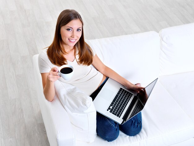 Happy young woman works on the laptop with cup of coffee on a sofa - high angle