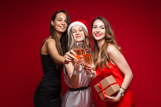 Happy young women friends with gift and champagne celebrating holiday together on party on red background, copy space