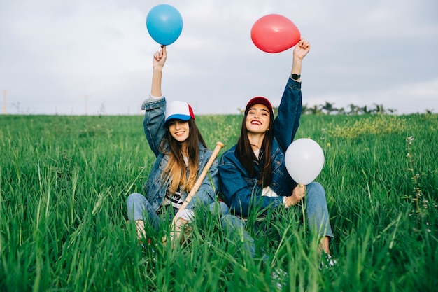 Happy young women with colored balloons in air sitting in field